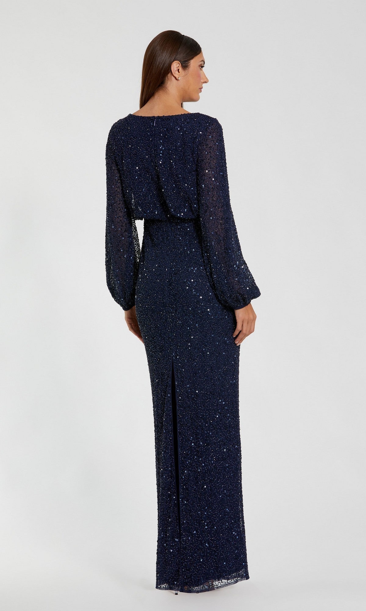 A woman is pictured standing and facing away from the camera against a plain background, wearing the Mac Duggal 94203 long formal dress. This navy blue gown features long sleeves, shimmering sequins, a belted waist, and a back slit. Its modest design creates an elegant silhouette ideal for sophisticated occasions.